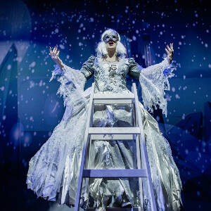 Grafika przedstawia kobietę w balowej sukni, białej peruce i masce na oczach. Kobieta stoi na drabinie z rozpostartymi rękami