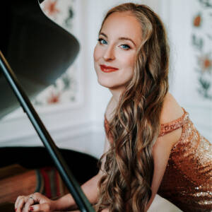 Zdjęcie przedstawia piękną, uśmiechniętą kobietę z długimi, kręconymi, jasnymi włosami, patrzącą w obiektyw, opartą o fortepian