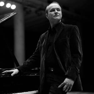 Zdjęcie czarno-białe pianisty Konrada Skolarskiego stojącego przy fortepianie i patrzącego w dal