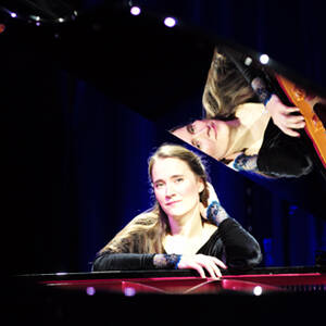 Pianistka siedzi w sali koncertowej przy fortepianie, patrzy prosto w obiektyw, w tle kotara oświetlona na granatowo