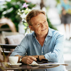 Michał Bajor siedzi uśmiechnięty przy stoliku z filiżanką kawy, trzyma przeciwsłoneczne okulary i gazetę