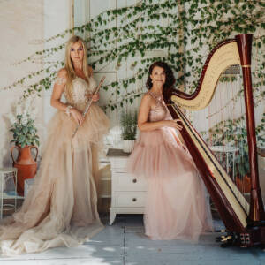 Zdjęcie przedstawia dwie kobiety ubrane w piękne ciemno różowe suknie.  Jedna trzyma fletm druga harfę.