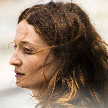 Zdjęcie portretowe kobiety. Kobieta patrzy w lewą stronę, ma rude włosy rozwiane przez wiatr. 