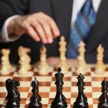 Grafika przedstawia drewnianą szachownicę, szachy oraz męską dłoń przestawiającą pionki 