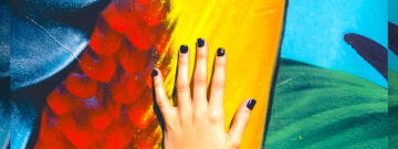 Grafika przedstawia kobiecą dłoń z pomalowanymi paznokciami na kolorowym obrazie